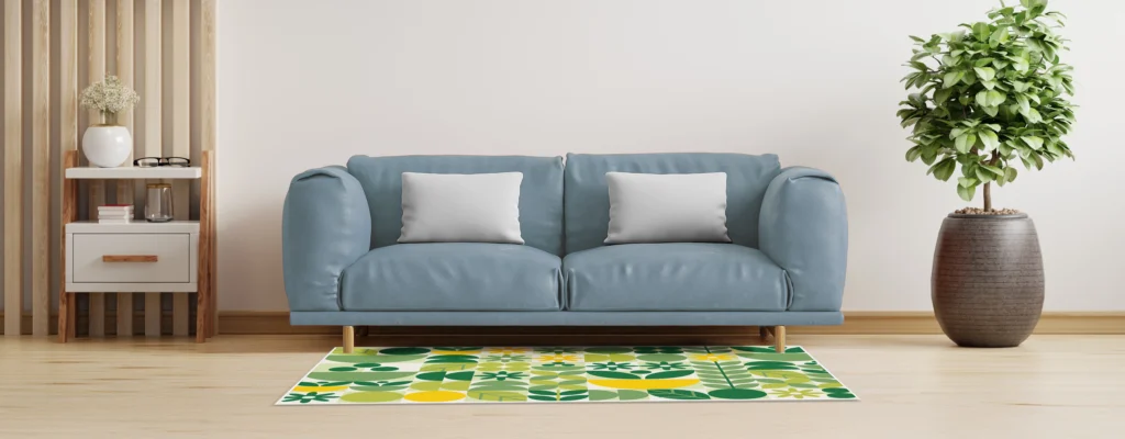 Blue sofa with two light-gray pillows in a bright living room, flanked by a wooden side table and a large potted plant; patterned green-yellow rug in front.