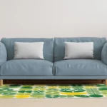 Blue sofa with two light-gray pillows in a bright living room, flanked by a wooden side table and a large potted plant; patterned green-yellow rug in front.