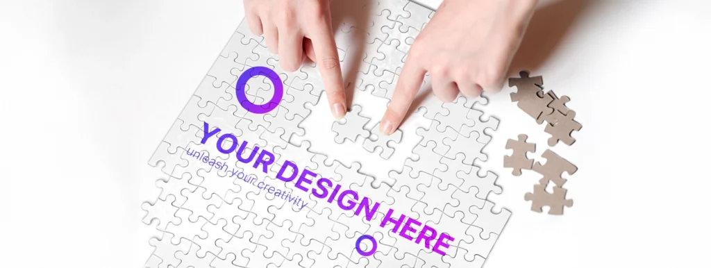 Hand placing a puzzle piece on a white jigsaw puzzle with the text 'YOUR DESIGN HERE' in purple and 'unleash your creativity'—conveying customizable design.