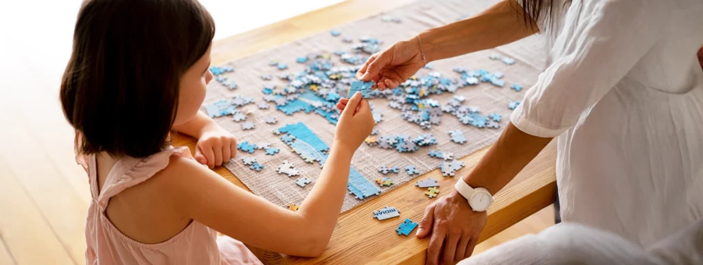 Child and adult placing blue puzzle pieces on a wooden table, working together on a jigsaw puzzle at home.