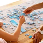 Child and adult placing blue puzzle pieces on a wooden table, working together on a jigsaw puzzle at home.