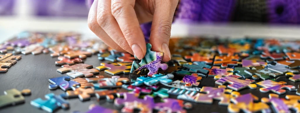 Person's hand placing a purple-green puzzle piece among colorful scattered pieces on a table