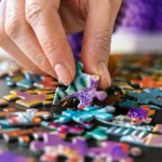 Person's hand placing a purple-green puzzle piece among colorful scattered pieces on a table