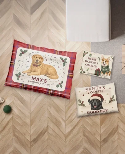 Dog-themed Christmas pillows on a light wood chevron floor: a red plaid pillow with a Golden Retriever and 'MAX'S CHRISTMAS NAP ZONE' plus a cream pillow reading 'MERRY WOOFMAS, CHARLIE!' with a corgi image; green ball nearby.