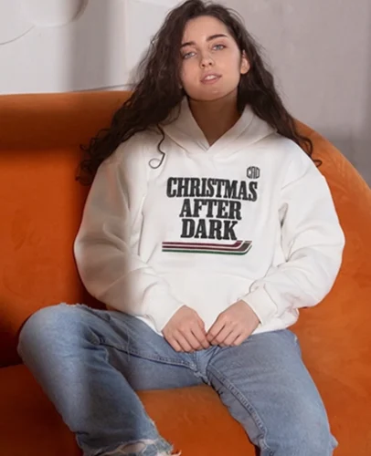 Young woman sitting on an orange couch wearing a white hoodie that reads 'CHRISTMAS AFTER DARK'.