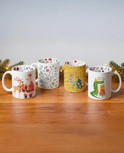 Group of white ceramic mugs with colorful holiday and floral designs arranged on a wooden table, with pine branch garland and warm fairy lights at the edges.