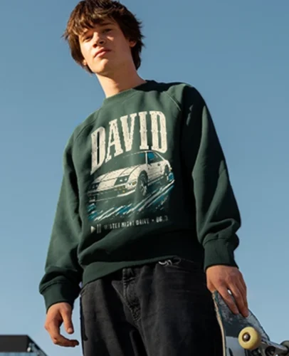 Young man wearing a dark green sweatshirt with 'DAVID' and a car graphic, holding a skateboard outdoors on a blue-sky day.