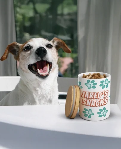 Smiling white and brown dog with mouth open sits at a table beside a Jared's Snacks cup of treats.
